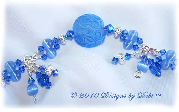 Designs by Debi Jewelry for Charity Piece for April 2010 to raise money for Autism Speaks. A one-of-a-kind artisan handmade bracelet with etched handmade glass lentil beads in a swirled pattern of shades of blue and a hint of violet, swarovski crystal sapphire and sapphire ab2x bicones, sapphire blue cat's eye beads, blue glass barrels, sterling silver twisted double curve and saucer beads and an abstract sterling silver toggle clasp OOAK.